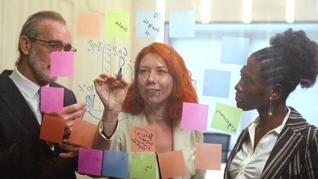 Smiling colleagues collaborate at a glass wall covered with colorful sticky notes and charts, brainstorming ideas and planning strategy during an office workshop meeting. 4k video