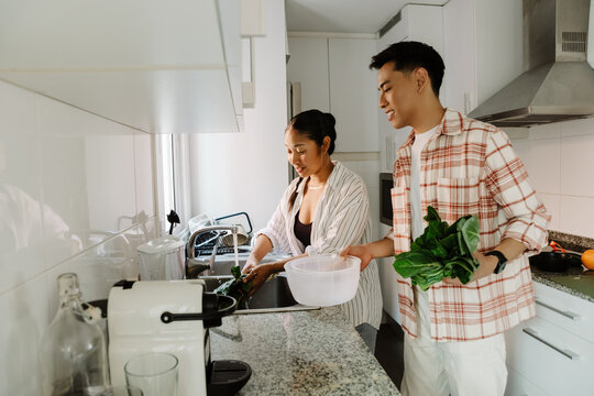 A man holds lettuce leaves and a colander while listening to a woman standing next to him washing the greens while they smile
