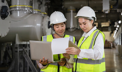 Two female engineers wearing safety helmets and reflective vests inspecting high-voltage gas-insulated switchgear (GIS) inside a power plant, teamwork, digital inspection industrial safety concept