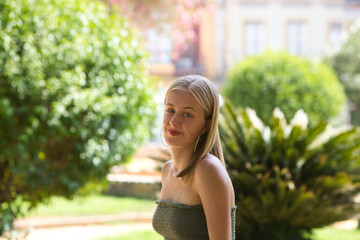 Portrait of a young, beautiful blonde woman wearing a green top, looking at the camera with a happy smile. The woman is in the park, sitting in the shade, on holiday in seville and sightseeing.