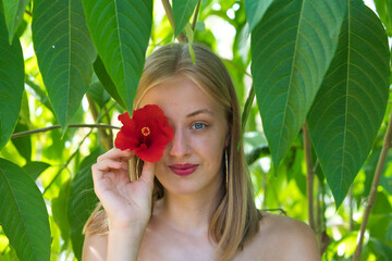 Portrait of a young, beautiful blonde woman with make-up and blue eyes, covering one eye with a red flower. The woman is surrounded by lush vegetation in the park and is smiling.