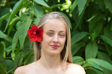 Portrait of a young, beautiful blonde woman wearing make-up and blue eyes with a red flower in her hair. The woman is surrounded by lush vegetation.