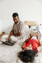 A woman lies on a bed and holds a book while looking at a laptop scrolled by a man sitting next to her