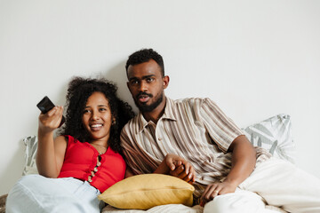 A man and a woman are talking while lying on a bed and looking ahead while she smiles and holds a remote control
