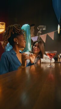 Vertical portrait shot of African American woman and her Caucasian friend recording video of celebrating birthday at bar counter