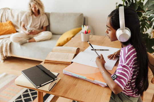 The daughter is sitting at the table and reading a book while holding a pencil and listening in headphones while her mother is sitting next to her on the couch