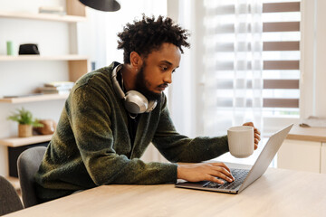 A male musician sits at a table and holds a cup while typing on a laptop