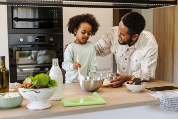 A father holds a bowl and hugs his daughter, who is holding a spoon, as they stand at the table