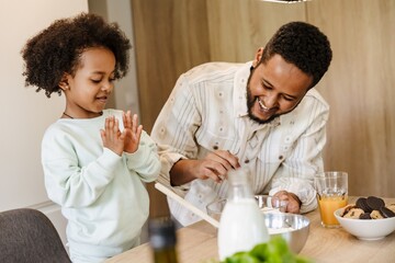 The daughter stands on a chair and applauds while watching her father cook and smiles while standing next to her