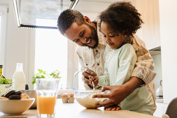 A father smiles and helps his daughter stir in a bowl