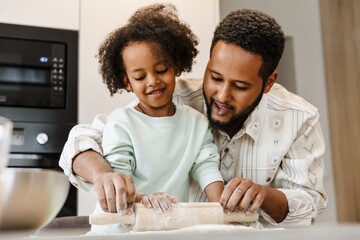 A father helps his daughter while they stand at the table and talk while they roll out the dough
