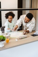 The father sprinkles the dough with flour while standing at the table next to his daughter