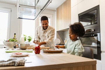 The daughter spreads the sauce on the dough while her father is standing next to her, holding a knife