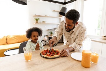 A father cuts a pizza while standing at a table with his daughter sitting at it