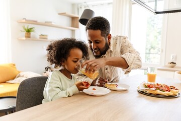 A father holds a glass from which his daughter drinks, who is sitting at the table and holding a piece of pizza