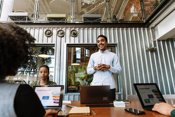 A male worker stands and talks while a group of three workers sit at a table next to him