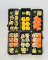 Assorted sushi rolls in black plastic takeout boxes, neatly arranged with ginger and wasabi on a white table, ideal for concepts of Japanese cuisine, delivery service, or modern healthy food lifestyle