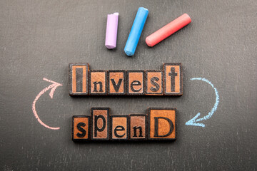 Words invest and spend made from vintage wooden letterpress blocks with chalk arrows on blackboard symbolizing financial decision making and money management concept
