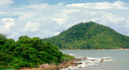 Tropical hill meets rocky Thai shoreline under a partly cloudy sky, with waves gently crashing—natural escape blending lush greenery, coastal terrain, and Southeast Asian atmosphere