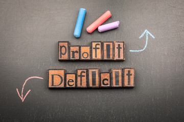 Words profit and deficit made from vintage wooden letterpress blocks with chalk arrows on blackboard symbolizing financial performance business balance and economic results
