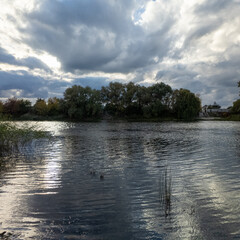 Calm river or lake with rippling water, surrounded by reeds and trees under dramatic cloudy sky. Sunlight breaks through clouds, creating a peaceful, natural autumn scene.