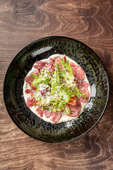 Gourmet beef carpaccio with shredded lettuce, grated cheese, and creamy dressing on a black plate. Elegant restaurant-style presentation on rustic wooden surface.