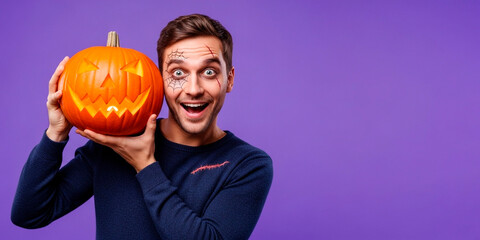 Man with Halloween makeup holding carved Jack-o'-lantern against violet background. Model in studio for Halloween celebration with copy space