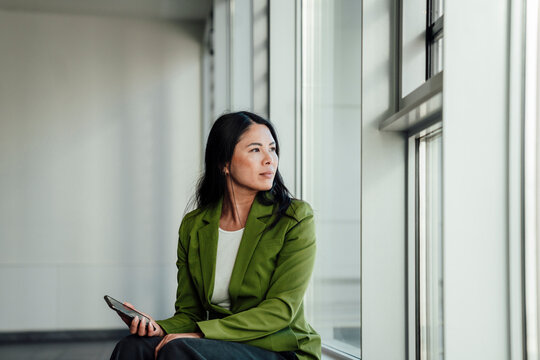 Businesswoman in green jacket working with smartphone by office window