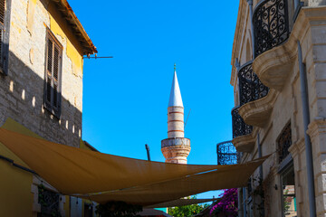 Minaret of Kebir Mosque in Limassol, Cyprus, as viewed from narrow street in  city old town. Traditional buildings with balconies and awnings line cobblestone alley, leading toward the mosque minaret