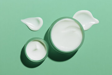 A minimalistic flat lay image featuring two clear jars of creamy moisturizer on a mint green background, with a couple of dollops of cream beside them.