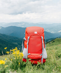 A bright red hiking backpack rests on green grass, surrounded by yellow wildflowers, against a panoramic mountain backdrop under a cloudy sky.