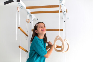 Smiling girl with long brown hair and turquoise T-shirt climbing a wooden gym ladder indoors with gymnastic rings. Concept of childhood, energy, fitness, healthy growth, and home exercise