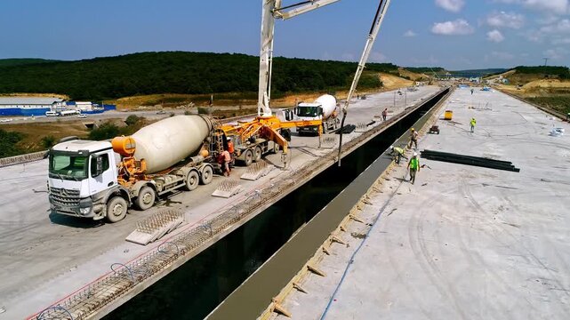 Drone aerial footage of concrete pouring on a highway bridge deck. Mixer trucks, concrete pump, and construction workers involved in large-scale infrastructure and engineering development.