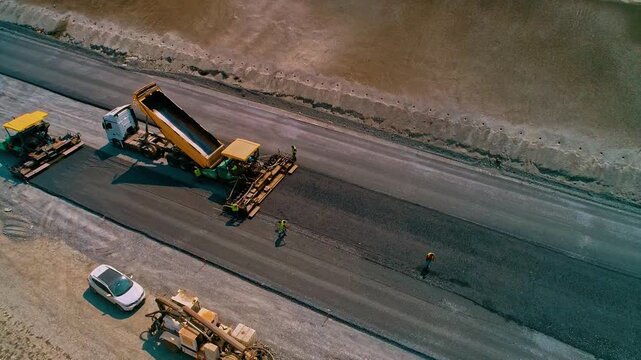 Drone footage of asphalt paving process with road construction machinery. Trucks, pavers, and rollers working on a modern highway infrastructure project.