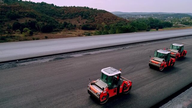 Drone footage of asphalt paving process with road construction machinery. Trucks, pavers, and rollers working on a modern highway infrastructure project.