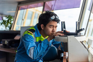 Young male officer in a blue and yellow uniform operates a navigational instrument on the bridge of...