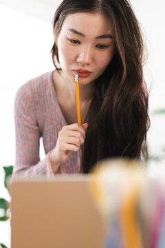 A young woman with long dark hair holds a pencil to her lips, deep in thought while looking at a laptop screen.