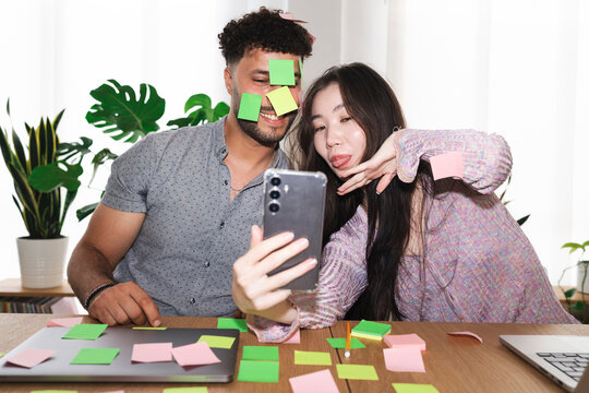 A man and woman playfully stick colorful notes on each other's faces while taking a selfie with a smartphone, surrounded by more notes and a laptop.
