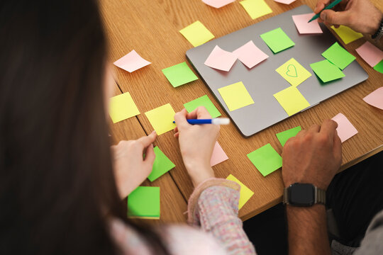 Two people collaborate on a project, drawing ideas on sticky notes scattered around a laptop on a wooden table.