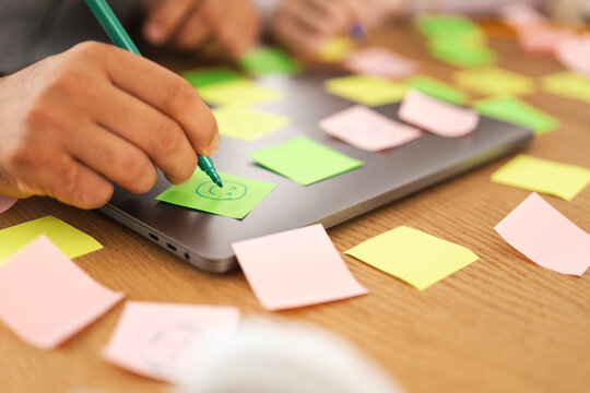 A person's hand uses a green marker to draw on a sticky note placed on a laptop, surrounded by other colorful notes on a wooden desk.