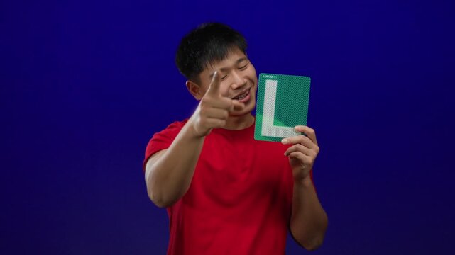 Young man in a red shirt points at camera holding an l sign over an isolated blue background wall.