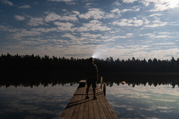 A man with a headlamp stands on a wooden pier in Estonia, looking at the beautiful cloudy night sky reflecting in the calm lake.