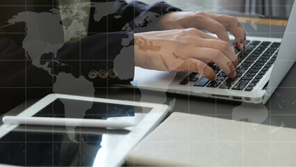 Businessman typing on laptop with digital tablet and notebook on desk, world map overlay symbolizes global communication and technology integration