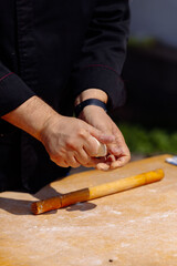 Chef's hands gently fold and prepare fresh, raw dough on a light wooden board, ready for cooking or baking in a rustic outdoor setting. 