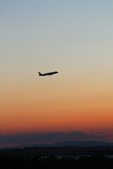 Paisaje, horizonte al atardecer en tonos degradados naranjas, con algo de nubes, se ve un avión cruzando el espacio visual, y el fondo montañoso como silueta, vertical