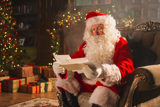 Merry Christmas. Happy Santa Claus reading letters from children in festive Christmas living room. Santa with xmas wish list letter on Christmas eve. Mail of Santa Claus. Winter Holiday Celebrations
