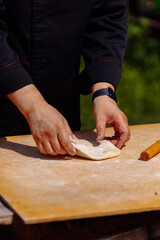 Chef's hands gently fold and prepare fresh, raw dough on a light wooden board, ready for cooking or baking in a rustic outdoor setting. 
