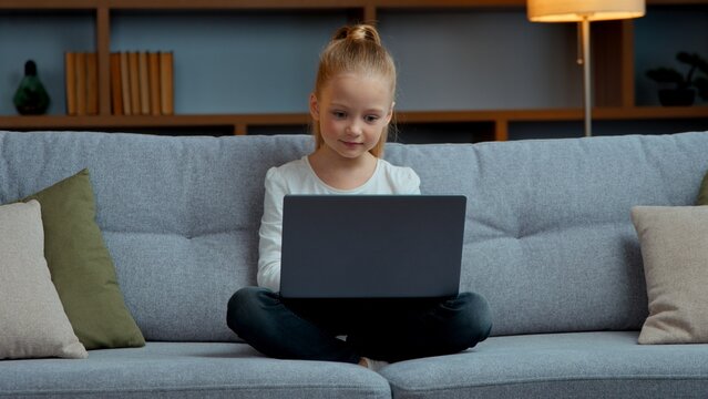 Caucasian little child girl using laptop on couch at home kid daughter with computer in living room modern technology and young generation schoolgirl playing online virtual games e-learning distant