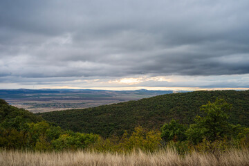 Mist drifts through a sunlit valley beneath forested hills and a dramatic, cloud-streaked sky.