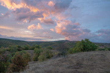 Pink, orange, and purple clouds drift over rolling hills at sunset, casting warm light across dry grass and distant trees.